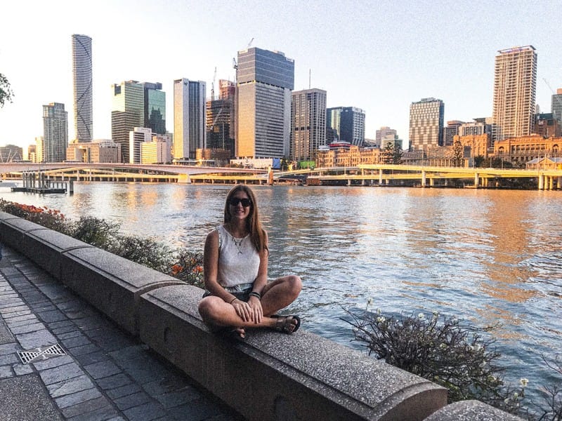 Chica morena sentada de rodillas en un muro con el fondo de la ciudad de Brisbane